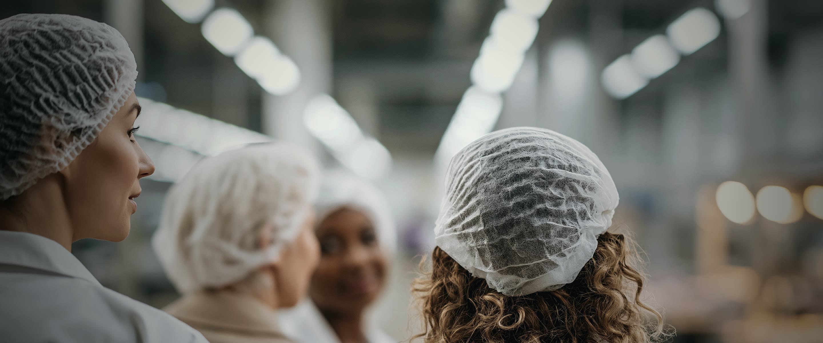 Four women in hairnets and white coats stand together on a processing floor, the plant's lit machinery stretching away out of focus behind them.