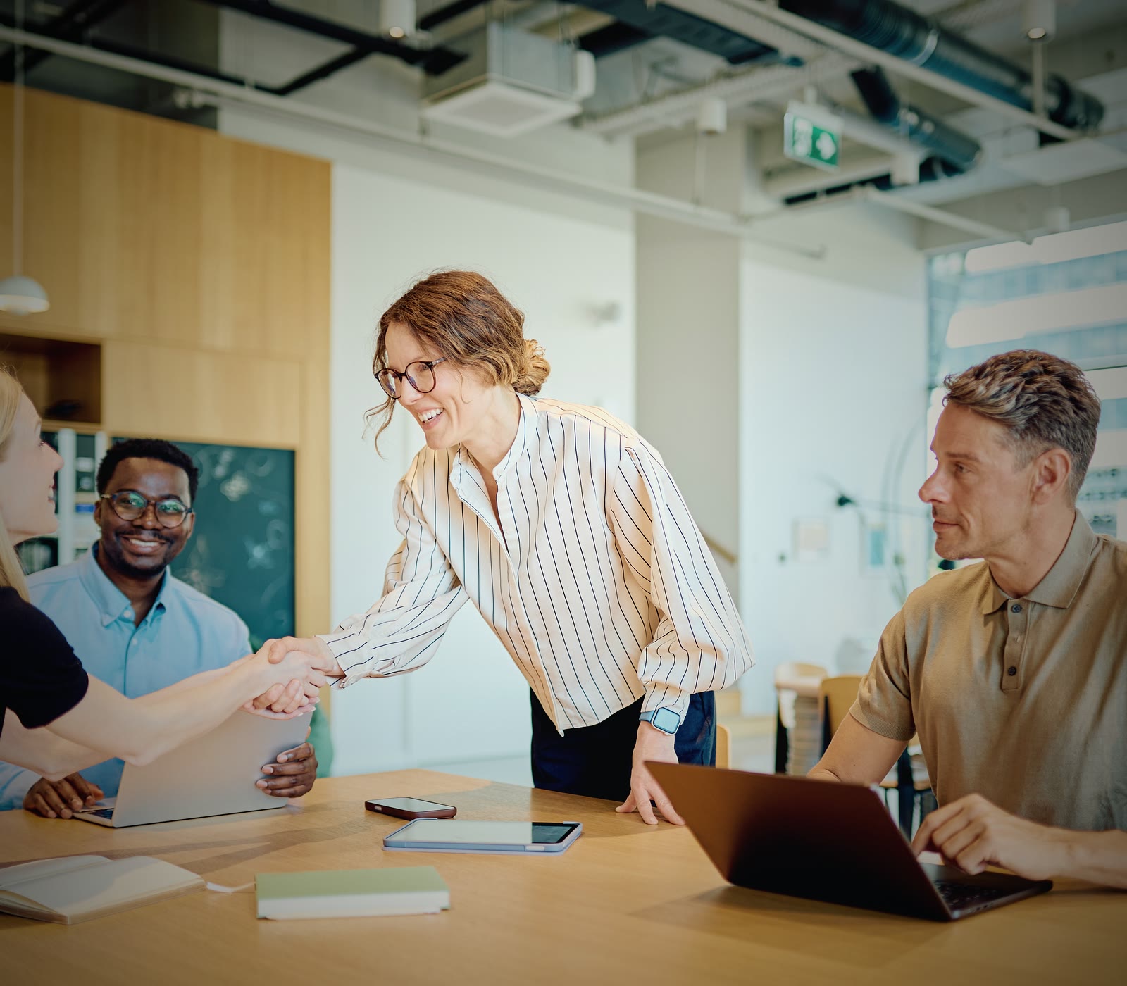 Two people shaking hands across a meeting table while two seated colleagues look on in a daylit office.