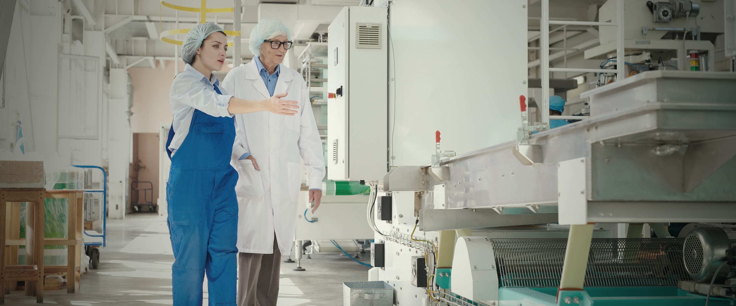 Two colleagues in white coats and hairnets stand at a machine on a plant floor, one gesturing towards it as they talk it through.
