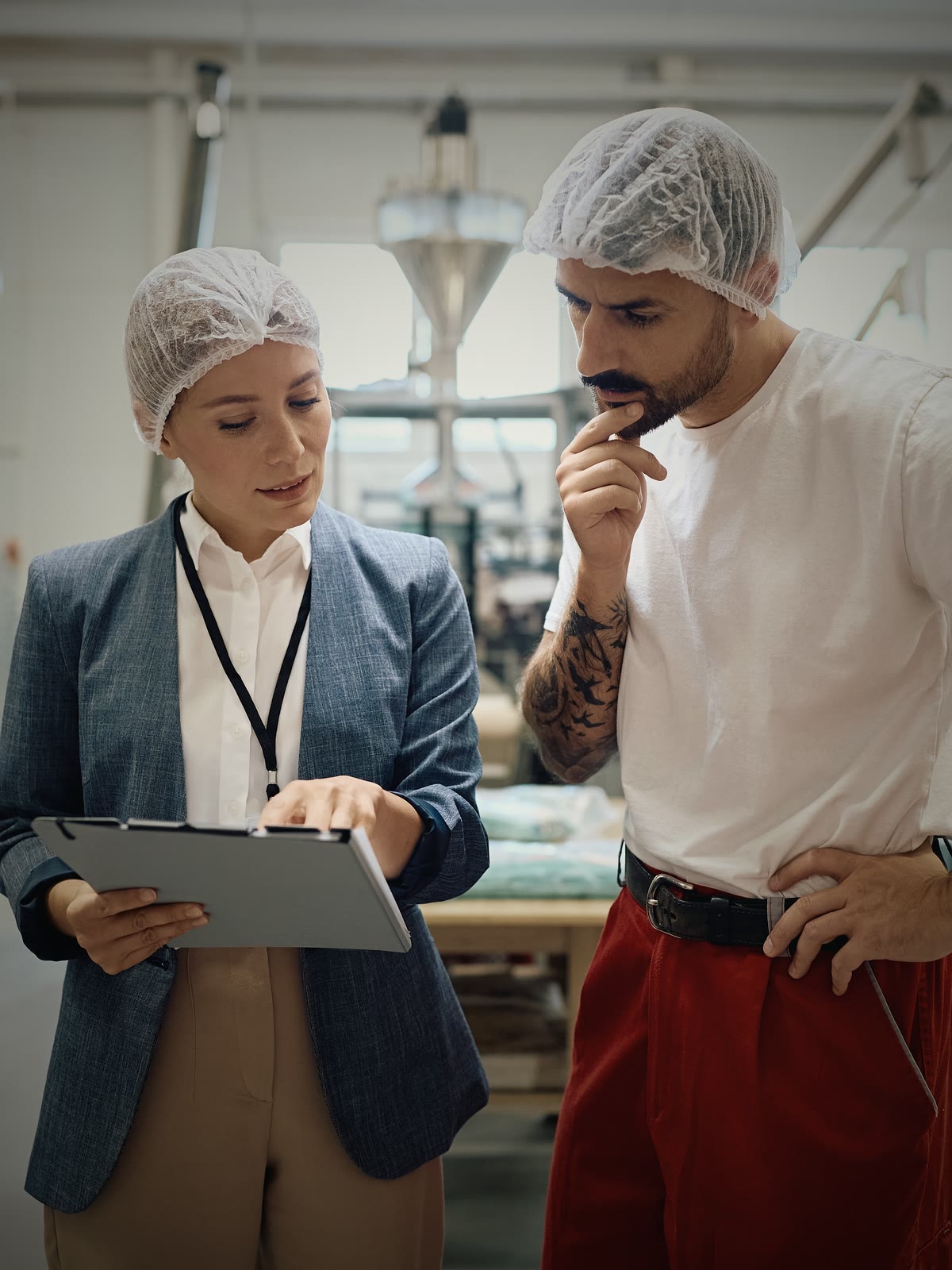 A woman in a blazer and hairnet holds out a clipboard to a worker in a hairnet and t-shirt on a food production floor, both reading it.