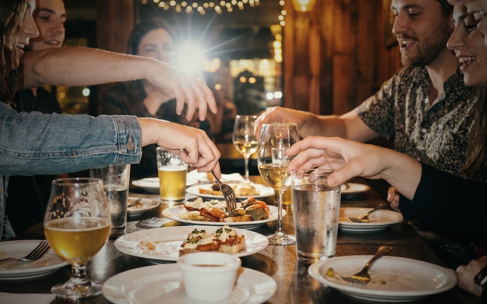 Guests leaning in around a restaurant table, sharing small plates under warm string lights.