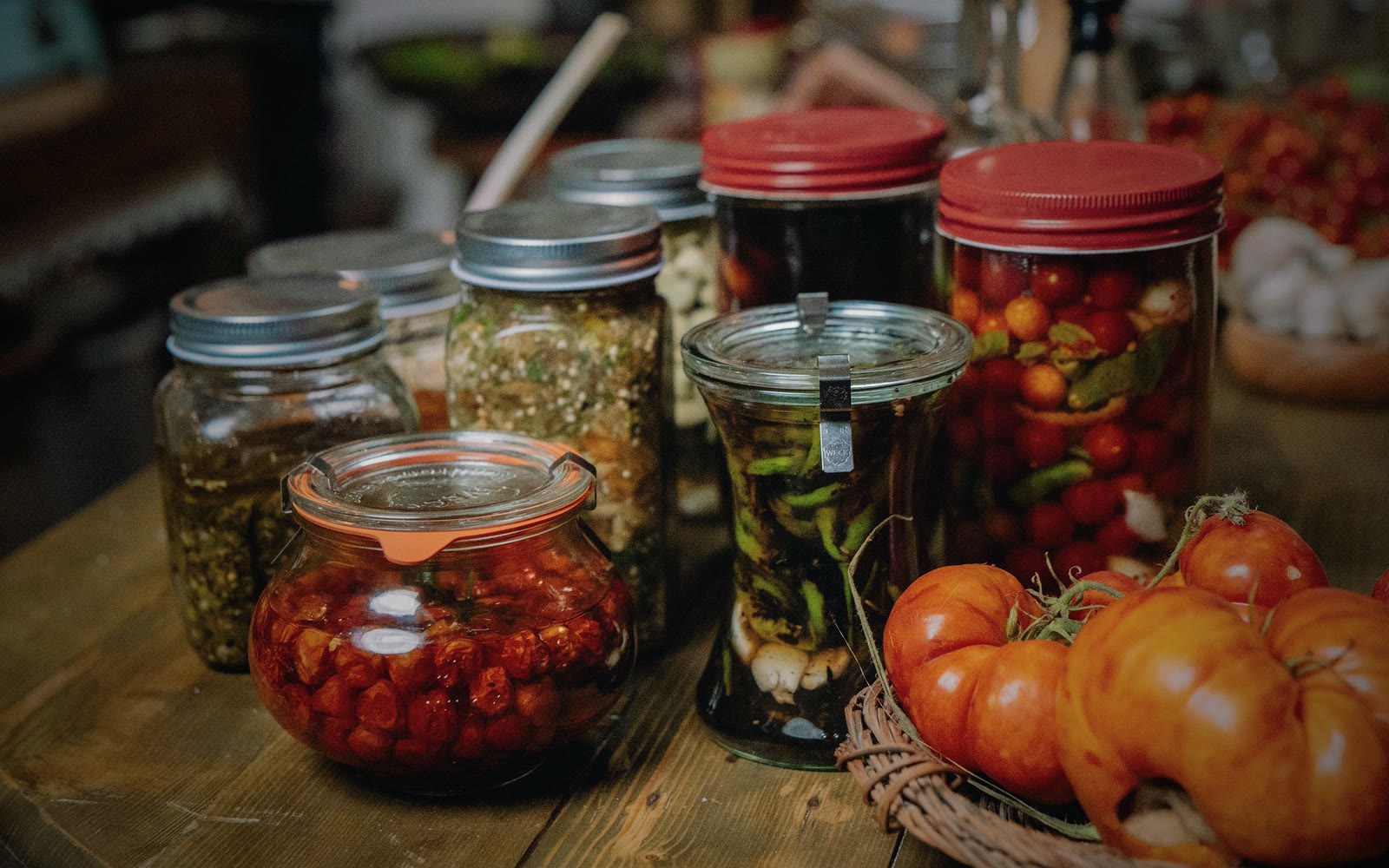 Glass jars of preserved vegetables and fresh tomatoes laid out on a wooden table